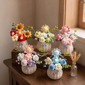 May include: Five small, round, matte beige ceramic pots, each containing a unique arrangement of crocheted flowers in various colors, including yellow, red, pink, blue, and white, accented with green fabric leaves and small white pearl beads.