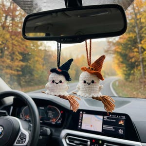 May include: Two crocheted ghost ornaments hanging from a rearview mirror. Each white ghost features a smiling face with pink cheeks and wears a pointed witch hat, one black and one orange. Each ghost is attached to a small brown yarn broomstick.