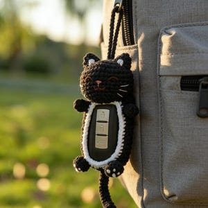 May include: A black and white crocheted cat key fob attached to a gray backpack. The cat-shaped cover has a smiling face, white accents, and a tail. The key fob is holding a car key.