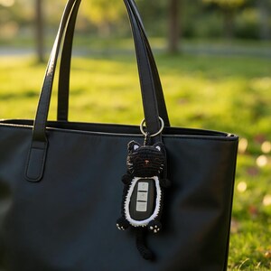 May include: A black crochet cat key fob cover featuring white accents around the face and belly, with a small tail and ears. The cat-shaped holder is attached to a silver metal ring and is currently hanging from the handle of a black leather tote bag.