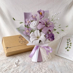 Puede incluir: Un ramo de flores de ganchillo con lirios morados y blancos, una rosa blanca y pequeñas flores moradas con tallos verdes. El arreglo está envuelto en papel lavanda y encaje blanco, atado con una cinta de color lavanda junto a una caja marrón.