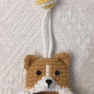 May include: A crocheted corgi-shaped item featuring tan and white yarn, black bead eyes, and a black stitched nose. A white yarn loop extends from the top, connected to a small, round, yellow and white striped crocheted ball ornament.