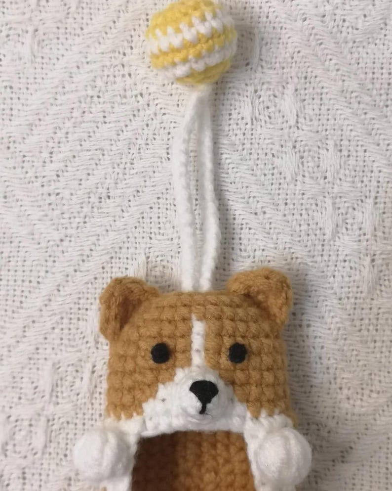 May include: A crocheted corgi-shaped item featuring tan and white yarn, black bead eyes, and a black stitched nose. A white yarn loop extends from the top, connected to a small, round, yellow and white striped crocheted ball ornament.