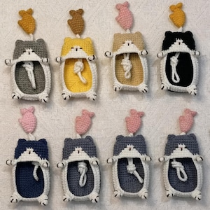 May include: Eight crocheted cat-shaped pouches arranged in two rows. Each pouch features a white trim, a small dangling fish charm in yellow, brown, or pink, and a white knotted cord inside. The cats come in gray, yellow, beige, black, and blue.