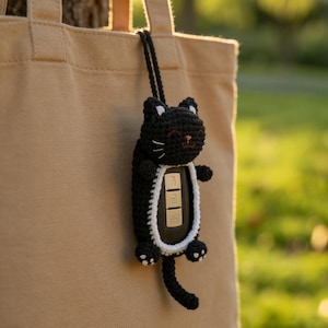 May include: A black crochet cat key fob cover featuring white inner ear details, white whiskers, and a white border around the central opening. The cat has small paws, a long tail, and is attached to a black cord for hanging from a beige tote bag.