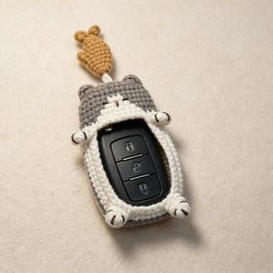 May include: A gray and white crocheted cat-shaped key fob cover featuring a small tan fish charm attached to the top. The cover encases a black rectangular car key remote with three buttons, showing the cat's ears, paws, and tail details.