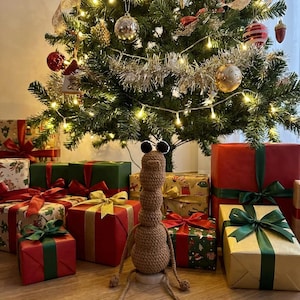 May include: A decorated Christmas tree with ornaments and lights, surrounded by wrapped gifts with ribbons. A brown crocheted creature with large eyes sits in front of the tree, adding a whimsical touch to the holiday scene.