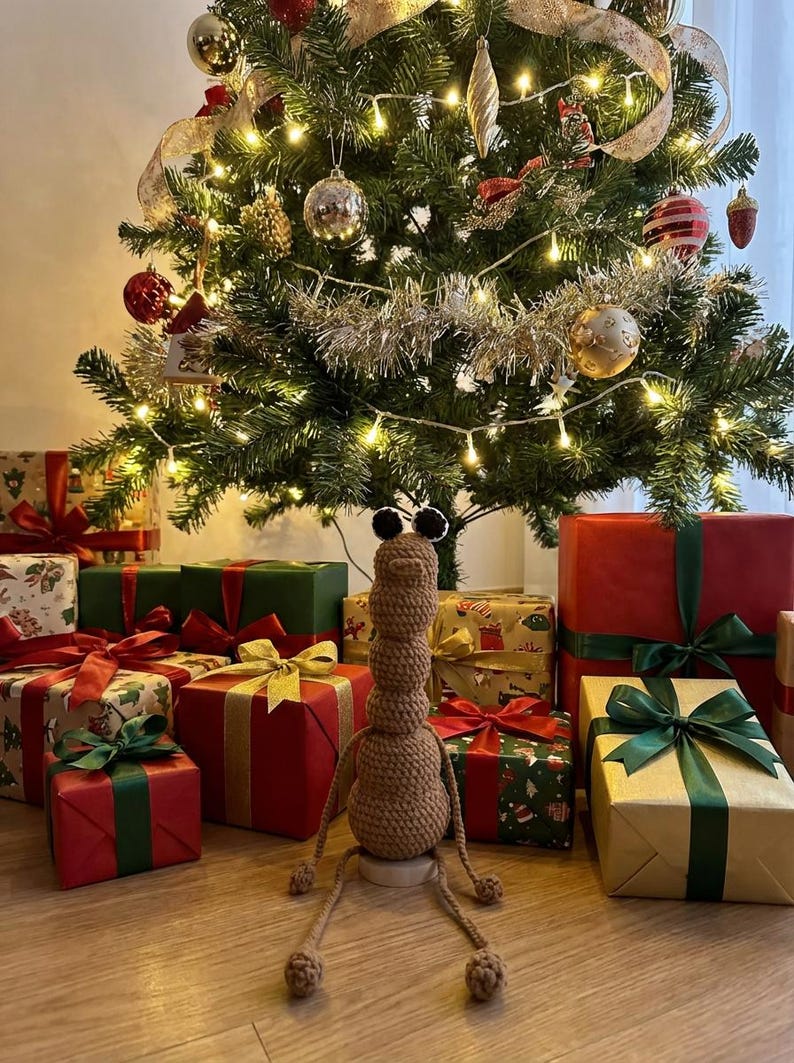 May include: A decorated Christmas tree with ornaments and lights, surrounded by wrapped gifts with ribbons. A brown crocheted creature with large eyes sits in front of the tree, adding a whimsical touch to the holiday scene.