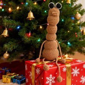 May include: A brown crocheted creature with black and white eyes sits atop a red gift box with white snowflake patterns and a gold ribbon. A Christmas tree with colorful lights and ornaments is in the background. Other wrapped gifts are at the base of the tree.