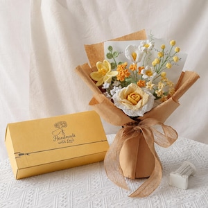May include: A crochet flower bouquet featuring yellow and white roses, daisies, and small blossoms wrapped in brown paper with a ribbon, accompanied by a yellow rectangular gift box labelled Handmade with Love and a small white cylindrical object.