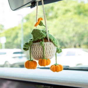 May include: Crocheted pumpkin car charm. The charm features a cream-colored pot with green leaves, an orange flower, and three small orange pumpkins hanging below. The charm is suspended by a cream-colored string, perfect for seasonal decor.
