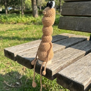 May include: A handmade, tan-colored crochet doll with a stacked body and dangling legs. The doll has a small head with a black and white accent. It is sitting on a wooden bench in a park setting.