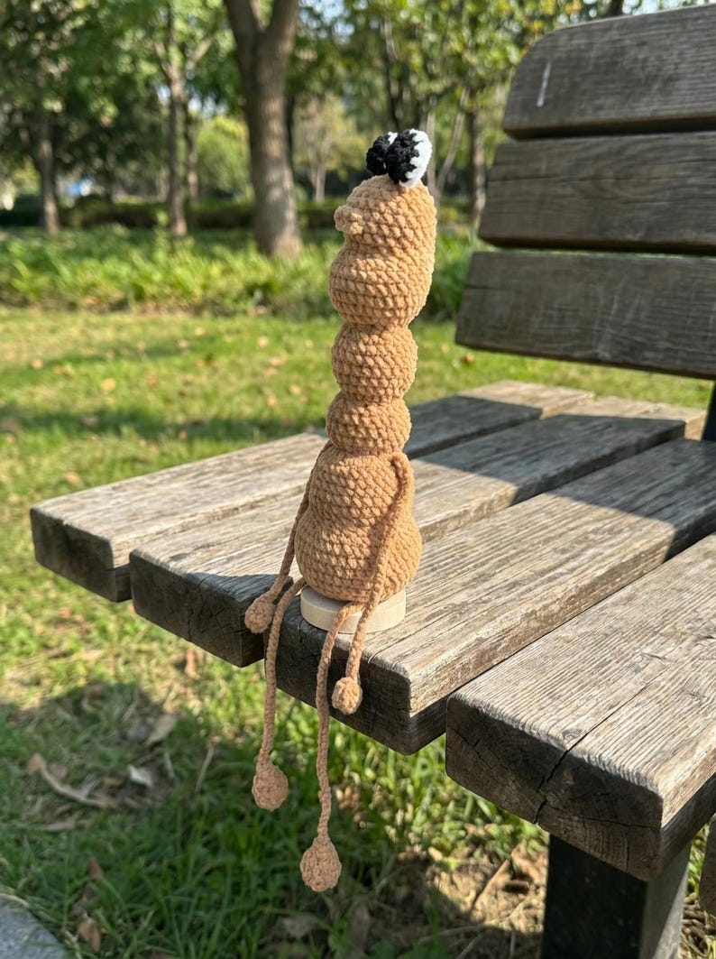 May include: A handmade, tan-colored crochet doll with a stacked body and dangling legs. The doll has a small head with a black and white accent. It is sitting on a wooden bench in a park setting.