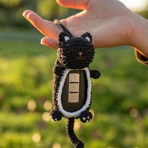 May include: A black crocheted cat-shaped key fob cover featuring white inner ear details, a white border around the central opening, and a long tail. The cover holds a black car key fob with three silver buttons visible through the center opening.