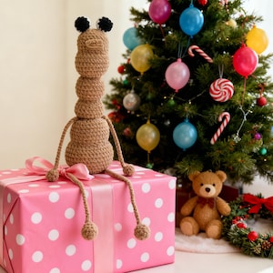 May include: A handmade crochet giraffe toy with black pompom antennae sits atop a pink polka dot gift box tied with a pink ribbon. A decorated Christmas tree with ornaments and a teddy bear are in the background.