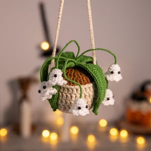 Puede incluir: Una maceta colgante de ganchillo hecha a mano con una maceta beige, tierra marrón y hojas verdes. Flores blancas con forma de fantasma con ojos negros cuelgan de la planta. La maceta está suspendida por una cuerda crema, ideal para Halloween.