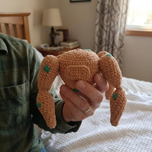 Peut inclure: Un jouet crocheté de couleur beige avec quatre membres articulés et un corps arrondi. Le jouet a de petits accents carrés verts sur les membres. Le jouet est tenu dans la main d'une personne.