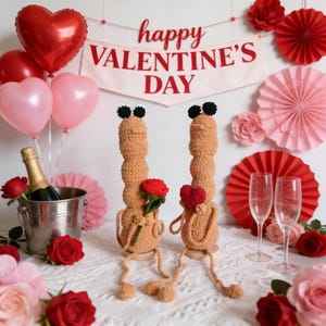May include: A Valentine's Day scene with two tan crochet figures holding a red rose and a heart. The background includes balloons, roses, champagne, and a banner that reads "happy VALENTINE'S DAY."