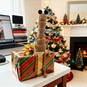 May include: A handmade, tan-colored crochet creature with black and white eyes sits atop a wrapped gift. The gift is wrapped in red and green striped paper with a gold bow. A Christmas tree with ornaments is in the background.