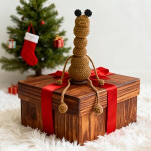 May include: A brown, crocheted insect-like figure sits atop a wooden gift box tied with a red ribbon. A small Christmas tree with ornaments and a stocking is in the background. The box is on a white, fluffy surface.