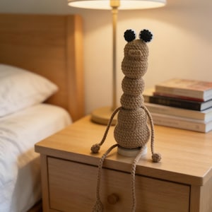 May include: A handmade, tan crochet caterpillar doll with black and white eyes and long, dangling legs. The caterpillar is sitting on a small, round, white base. The doll is on a wooden nightstand next to a lamp and a stack of books.
