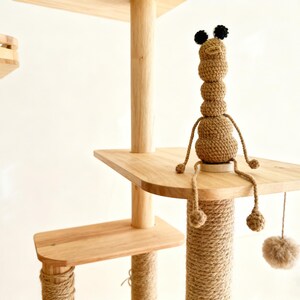May include: A multi-level cat tree with wooden platforms and posts wrapped in natural rope. A handmade, brown crochet worm-like toy with black pompom ears sits on a platform. A dangling pom-pom toy hangs below.