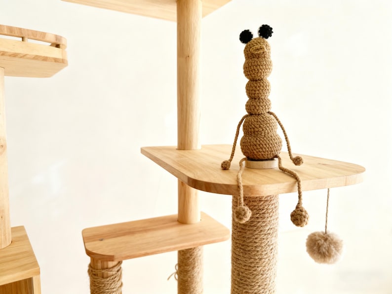 May include: A multi-level cat tree with wooden platforms and posts wrapped in natural rope. A handmade, brown crochet worm-like toy with black pompom ears sits on a platform. A dangling pom-pom toy hangs below.