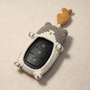 May include: A gray and white crocheted cat-shaped key fob cover featuring a small brown crocheted fish attached to the top. The cover surrounds a black car key remote with three buttons, displaying a lock, unlock, and trunk release icon.