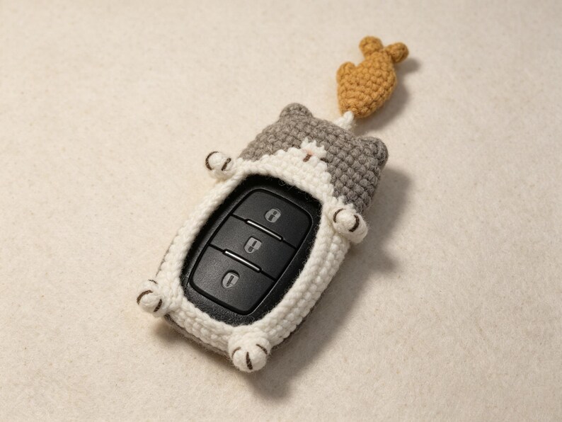 May include: A gray and white crocheted cat-shaped key fob cover featuring a small brown crocheted fish attached to the top. The cover surrounds a black car key remote with three buttons, displaying a lock, unlock, and trunk release icon.