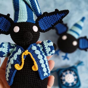 Op de afbeelding: Een zwarte gehaakte vleermuispop met grote oren, met een blauw en lichtblauw gestreepte puntmuts en een bijpassend blauw, lichtblauw en wit patroonjasje met een geel strikje. De vleermuis heeft twee ronde witte ogen en een zwarte neus.