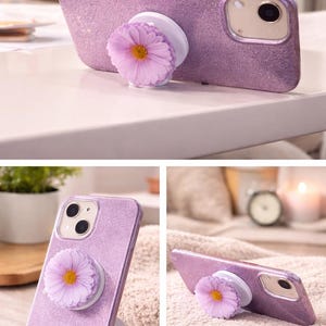 May include: A lavender glitter phone case with a daisy-shaped phone grip. The phone case has a camera and flash. The daisy grip is light purple with a yellow center. The phone case is on a white surface.