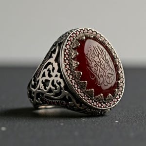 May include: A silver ring with an oval, deep red stone. The stone is engraved with Arabic script and surrounded by small, red gemstones. The ring has an ornate, filigree design on the band.