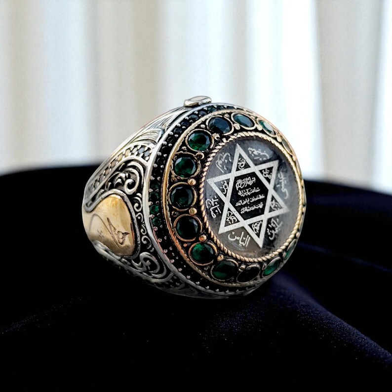 May include: A silver ring with a gold accent and a clear, circular center. The center features a six-pointed star and text. The ring is adorned with green gemstones and intricate detailing. The ring is set against a dark background.