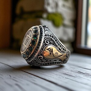 May include: A detailed silver ring with gold accents and a large oval stone. The ring features intricate scrollwork, black and green gemstones, and a gold-colored emblem. The ring is displayed on a wooden surface.