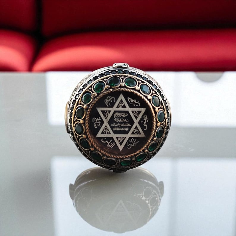 May include: A detailed, ornate ring with a central Star of David design. The ring features a dark background with gold-colored text and a surrounding border of green gemstones. The ring's band is a gold color with a dark background.