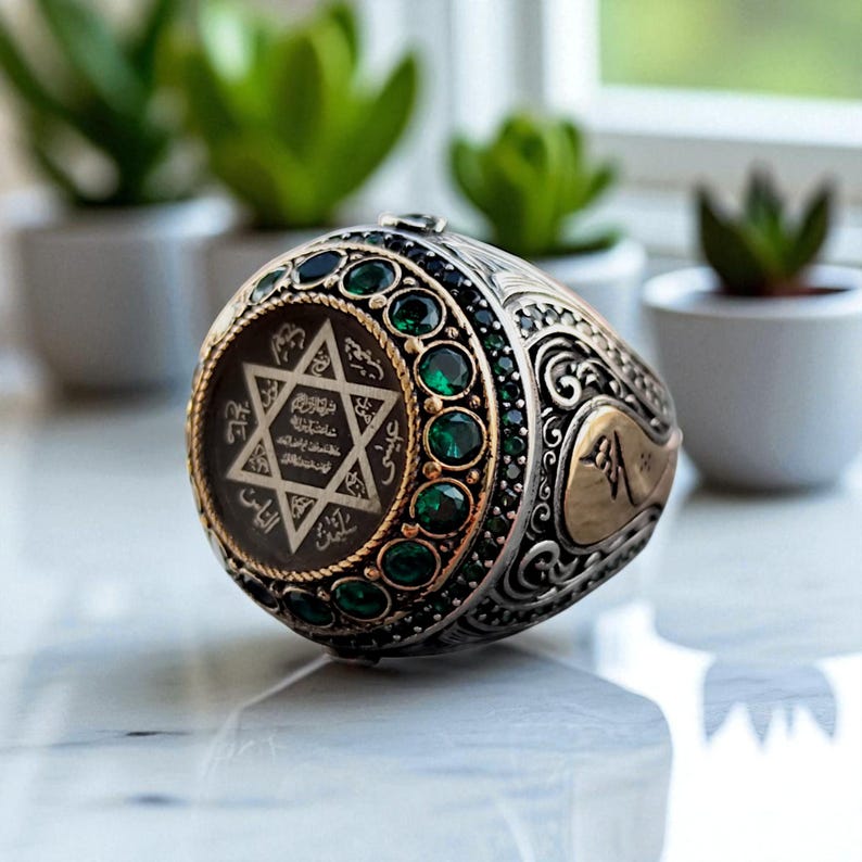 May include: A silver ring with a detailed design, featuring a central emblem with a star and text. The ring is accented with green gemstones and gold-colored accents. The ring is displayed on a white surface.