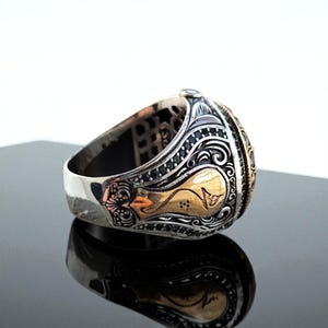 May include: A silver ring with gold accents and intricate detailing. The ring features a central design with a gold-colored emblem and is adorned with small, dark green stones. The band has a polished finish, reflecting the light.