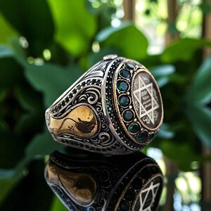 May include: Silver ring with gold accents and a central emblem featuring a Star of David. The ring is adorned with green gemstones and intricate scrollwork. The ring is displayed on a reflective surface.