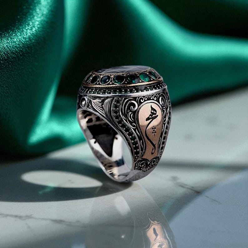 May include: A silver ring with an oval-shaped top featuring a dark green gemstone and smaller green stones. The ring has intricate detailing, including black and gold accents, and a gold-colored emblem on the side.
