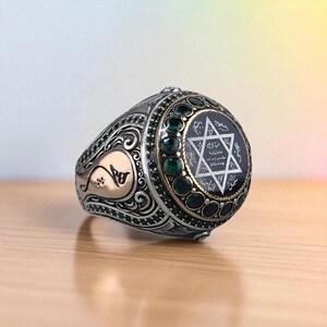 May include: Silver-toned ring with intricate filigree and gold accents. The ring features a black center stone with a white Star of David design and surrounding green gemstones. The ring is set on a wooden surface.