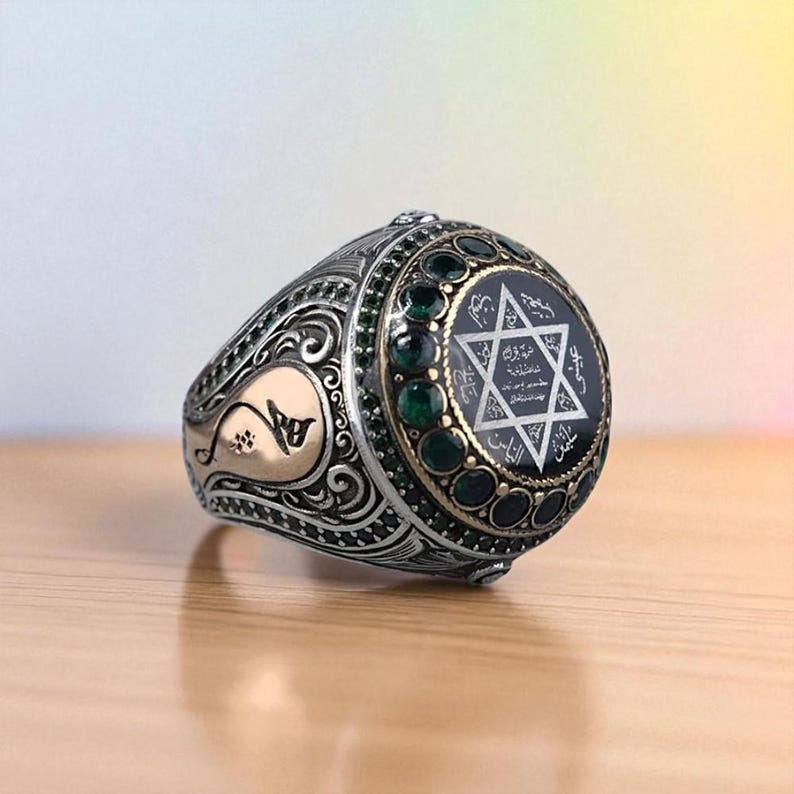 May include: Silver-toned ring with intricate filigree and gold accents. The ring features a black center stone with a white Star of David design and surrounding green gemstones. The ring is set on a wooden surface.