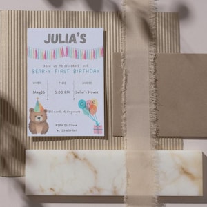 Może przedstawiać: Wyświetlana jest zaproszenie urodzinowe z tekstem "JULIA'S BEAR-Y FIRST BIRTHDAY". Zaproszenie zawiera ilustrację niedźwiedzia, balony i prezent. Zaproszenie znajduje się na teksturowanym tle z wstążką i brązową kopertą.