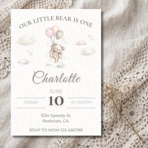 Puede incluir: Una tarjeta de invitación de color claro con el texto "OUR LITTLE BEAR IS ONE" en la parte superior. La tarjeta presenta un oso de peluche sosteniendo globos, el nombre "Charlotte" y detalles del evento para el 10 de junio a las 2:00 PM. Incluye dirección e información RSVP.