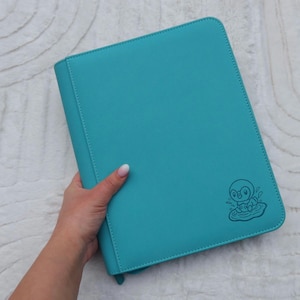 Może przedstawiać: Turkusowa teczka z zapięciem na zamek błyskawiczny. Mała, ciemnoniebieska ilustracja postaci z kreskówki znajduje się w prawym dolnym rogu. Teczka trzymana jest w dłoni.