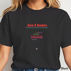 Könnte beinhalten: Schwarzes T-Shirt mit dem Text "Gen X Gamer" in Rot, über einer pixeligen Grafik eines Raumschiffs und außerirdischer Invasoren. Das Design enthält grüne, rote und blaue pixelierten Elemente, die an Retro-Videospiele erinnern.