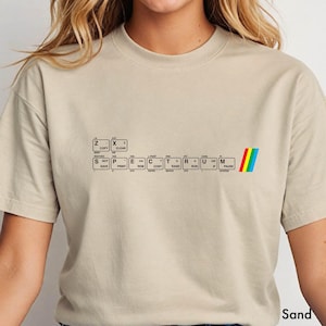 Puede incluir: Camiseta color arena con un diseño inspirado en un teclado que deletrea la palabra "SPECTRUM" con teclas. Una franja de arcoíris está a la derecha. La camiseta está hecha de un material suave. La palabra "Sand" está abajo a la derecha.
