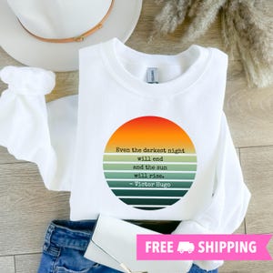 May include: White sweatshirt with a circular graphic featuring an orange and green sunset design. The graphic includes the quote "Even the darkest night will end and the sun will rise. - Victor Hugo." The sweatshirt is paired with blue jeans and a white clutch.