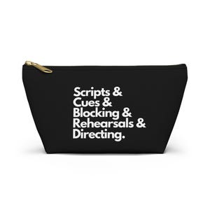 May include: Black zippered pouch with the text "Scripts & Cues & Blocking & Rehearsals & Directing" in white. The pouch has a gold-colored zipper and a trapezoid shape.