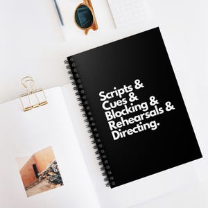 Puede incluir: Un cuaderno negro con espiral con texto blanco que dice "Scripts & Cues & Blocking & Rehearsals & Directing." El cuaderno está abierto sobre una superficie blanca con un clip dorado y gafas de sol.