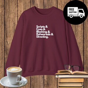 Könnte beinhalten: Ein weinrotes Sweatshirt mit dem weißen Text "Scripts & Cues & Blocking & Rehearsals & Directing". Eine Kaffeetasse, eine Brille und ein Buch befinden sich im Vordergrund. Ein schwarzer Kreis mit einem weißen LKW und den Worten "FREE SHIPPING" befindet sich im Hintergrund.